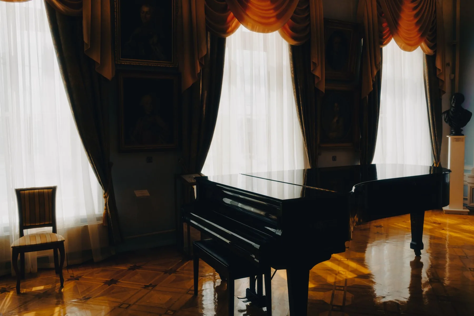 A black grand piano on a parquet floor beside tall draped windows in an old drawing room